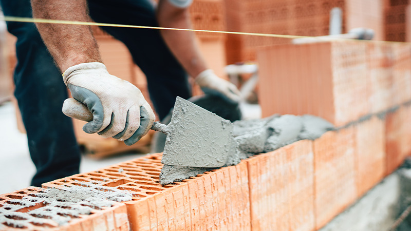 person applying mortar with a trowel on a brick wall during construction working on a masonry project with six bricks in place